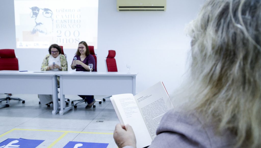 Fotografia registra uma pessoa de costa manuseando um livro. Ao fundo, é possível identificar uma projeção no quadro que traz o txto: TRIBUTO A CAMILO CASTELO BRANCO - 200 ANOS.