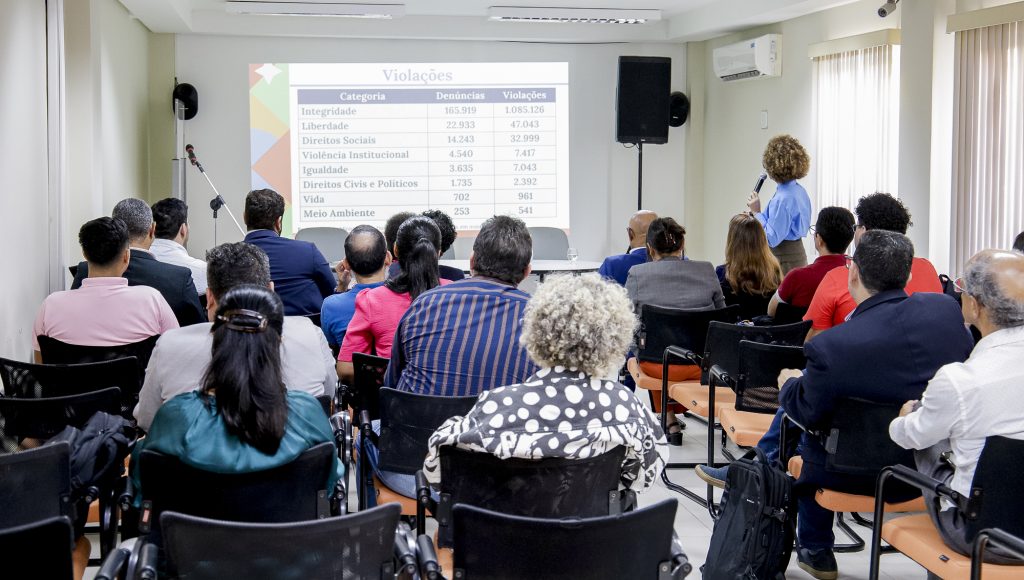 Fotografia mostra várias pessoas de costas, elas olham para uma apresentação projetada por um data show, na parede, onde é possível identificar a palavra "Violações". Na frente, uma das pessoas está de pé e fala ao microfone, olhando para a apresentação.