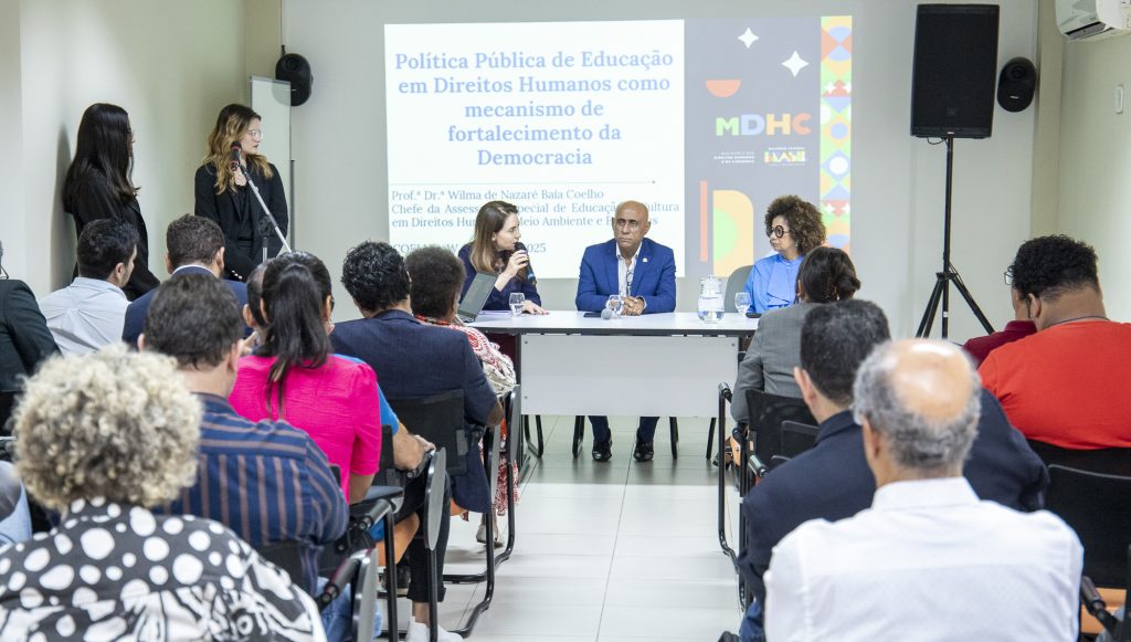 fotografia mostra várias pessoas em uma sala. Algumas estão sentadas em cadeiras, de costas. No fundo da sala, estão o reitor da e a vice reitora da UFPA, ao lado dA PROFESSORA wILMA bAÍA, sentados, atrás de uma mesa. Ao fundo, uma apresentação é projetada na parede.