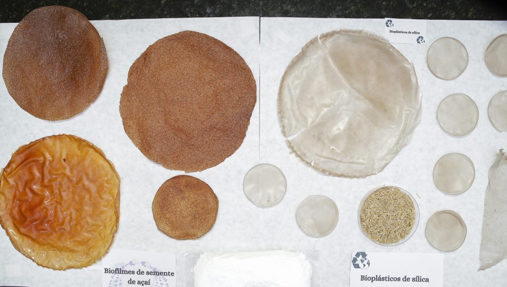 Fotografia de círculos em diferentes tons de marrom, dispostos sobre um papel branco. No papel branco há a identificação: BIOFILMES DE SEMENTE DE AÇAÍ e BIOPLÁSTICOS DE SÍLICA.