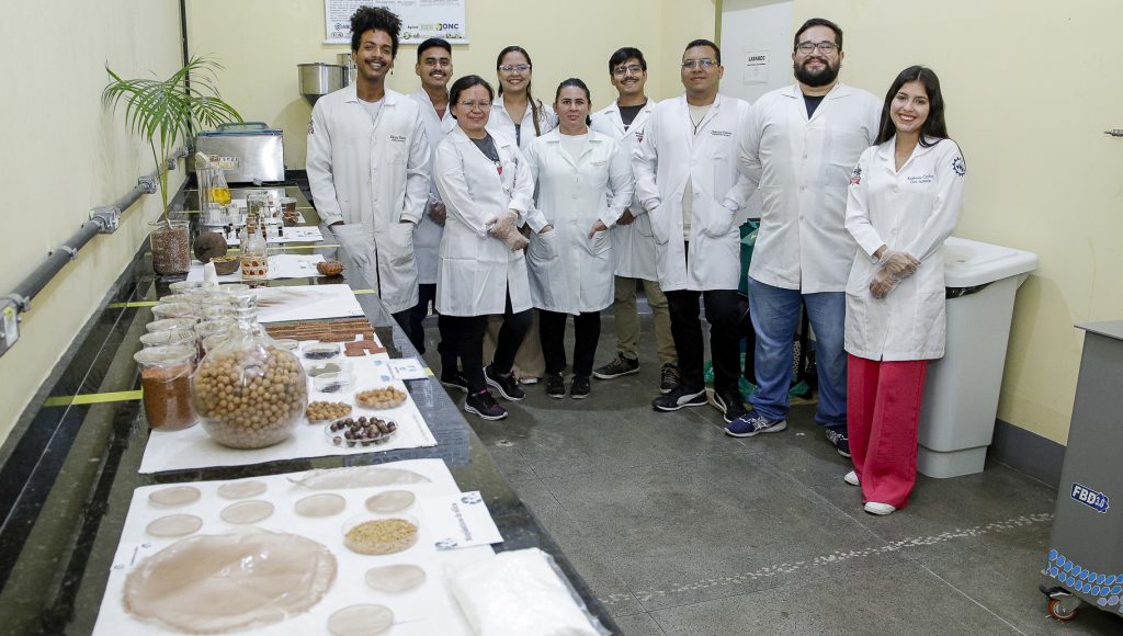 FOTOGRAFIA POSADA DE UM GRUPO DE PESSOAS DENTRO DE UM LABORATÓRIO COM DIVERSOS PRODUTOS DISPOSTOS EM UMA BANCADA. tODAS AS PESSOAS VESTEM JALECOS BRANCOS.