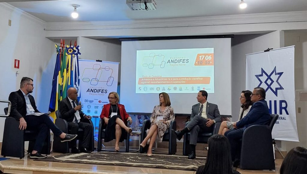 Fotografia registra seis pessoas sentadas em cadeiras, a frente de um grande telão. Entre as pessoas está o reitor da UFPA, Gilmar Pereira da Silva.