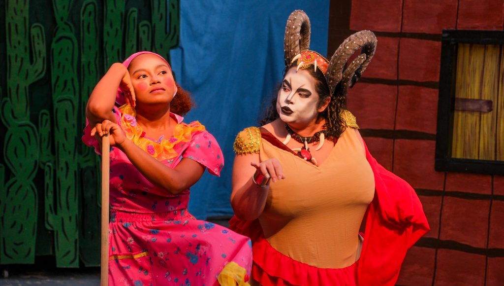 Fotografia registra duas mulheres contracenando em um teatro. Uma está apoiada em uma vassoura e a outras gesticula ao falar.