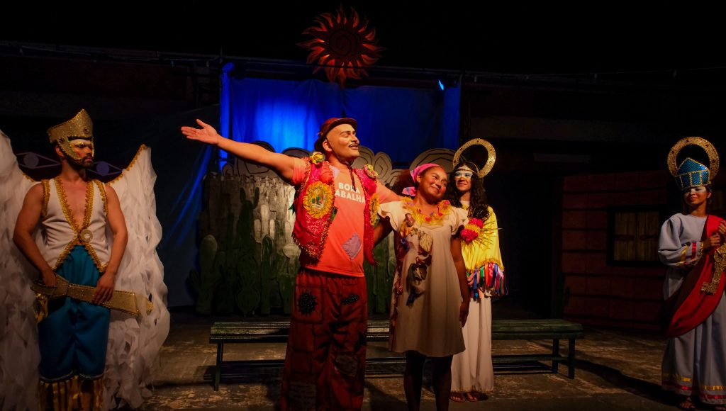 Fotografia de uma cena de teatro. Nela estão presentes um grupo de atores. Em destaca, um homem e uma mulher sorriem abraçados.