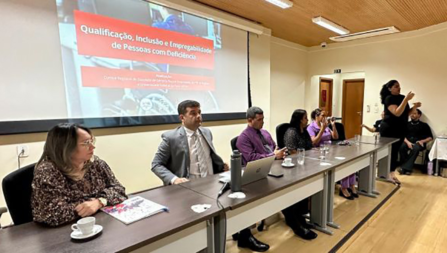 Fotografia registra a mesa de um evento composta por cinco pessoas. Na lateral da mesa, uma pessoa, vestida de preto, gesticula em movimento de LIBRAS. Ao fundo da mesa um projeção destaca o texto: QUALIFICAÇÃO, INCLUSÃO E EMPREGABILIDADE DE PESSOAS COM DEFICIÊNCIA.