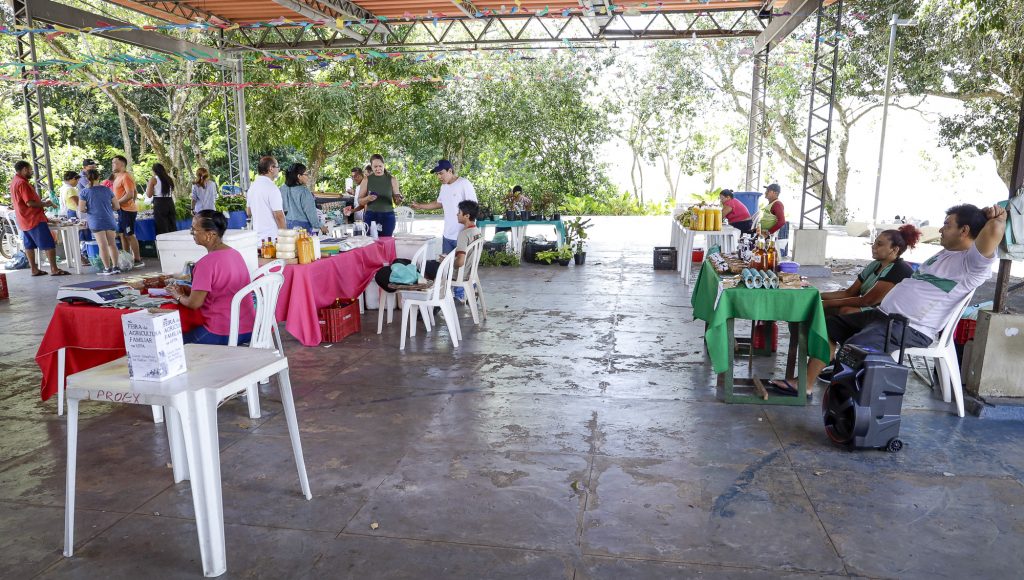 Fotografia registra um amplo espaço com várias mesas dispostas. Uma expõe garrafas de mel, outra queijos e pimenta, outra garrafas com tucupi e uma quarta dispõe de plantas em vasos.