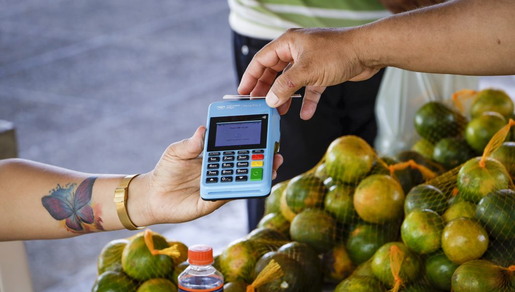 Fotografia registra uma mão que segura um cartão aproximando-o de uma máquina azul. Ao fundo, podemos ver algumas laranjas.