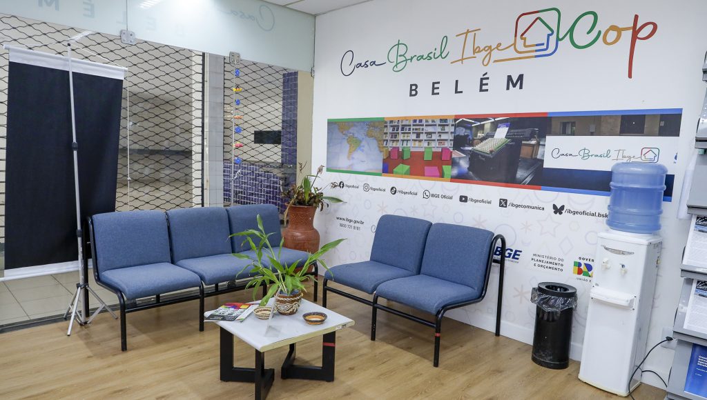 Fotografia de um espaço interno de uma sala, em destaque, no topo da imagem há uma arte na parede, seguida do texto: CASA BRASIL IBGE - COP - BELÉM.
