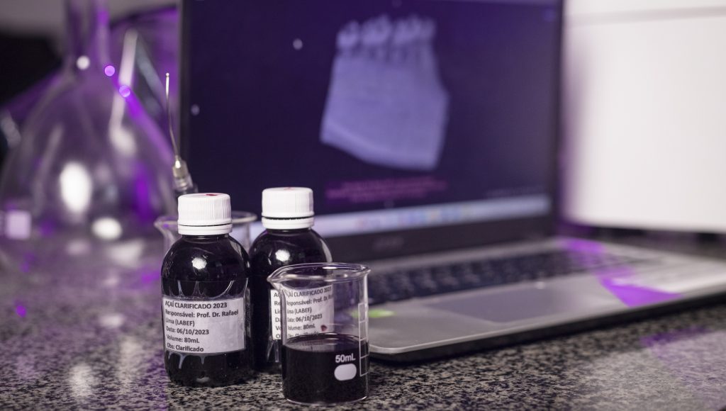 Fotografia exibe uma composição de objetos em uma bancada. Em destaque, dois frascos escuro com rótulo "AÇAÍ CLARIFICADO 2023", indicando um produto de açaí com tratamento específico. Ao fundo, um laptop.
