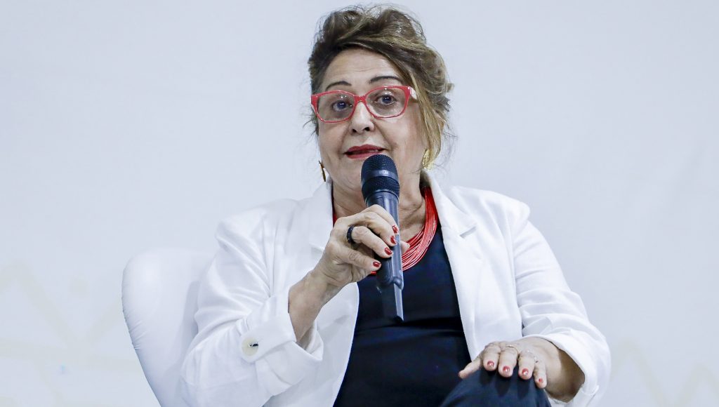 Fotografia registra a professora Edna Castro, enquanto ela fala ao microfone.