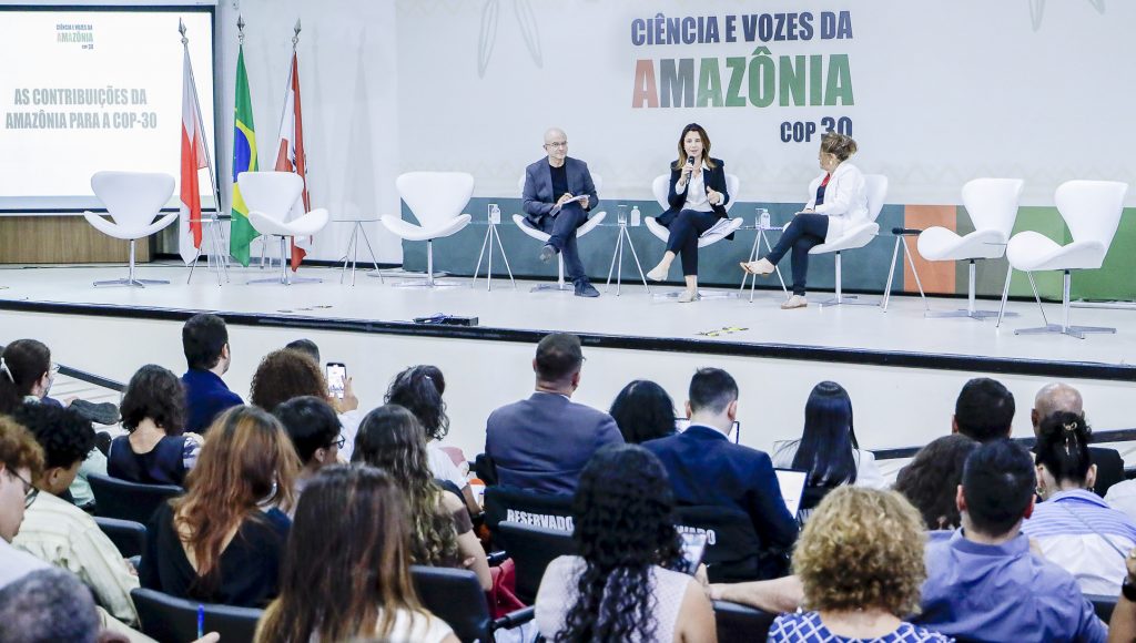 Fotografia registra parte de uma plateia de costas. Ao fundo, é possível identificar três pessoas sentadas em cadeiras dispostas a frente de um painel com o seguinte texto: CIÊNCIAS E VOZES DA AMAZÔNIA COP 30
