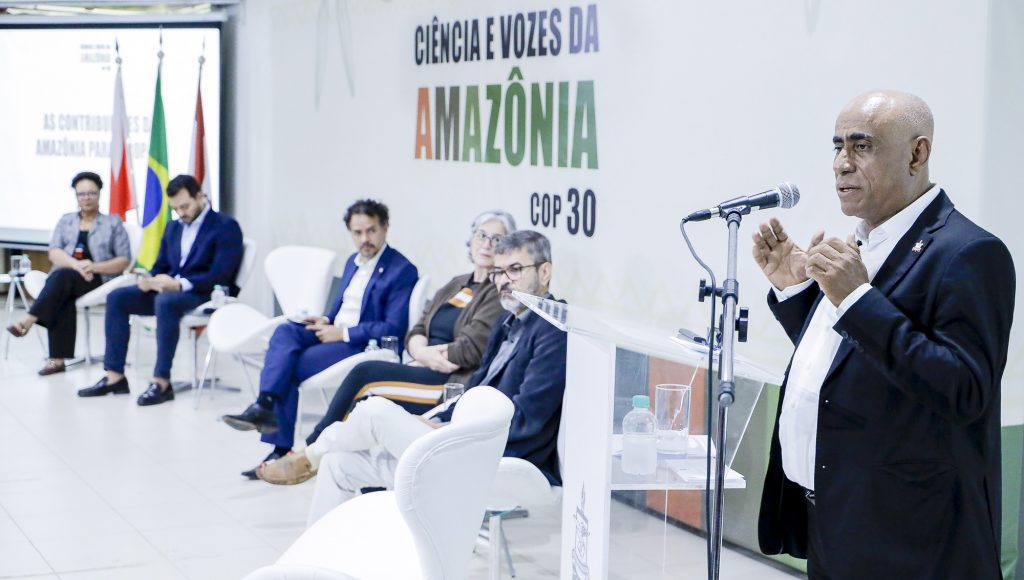 Fotografia registra o reitor da UFPA, Gilmar Pereira da Silva, falando ao microfone. Em segundo plano, estão quatro pessoas sentadas em cadeiras disposta a frente de um painel com o seguinte texto: CIÊNCIA E VOZES DA AMAZÔNIA COP 30.