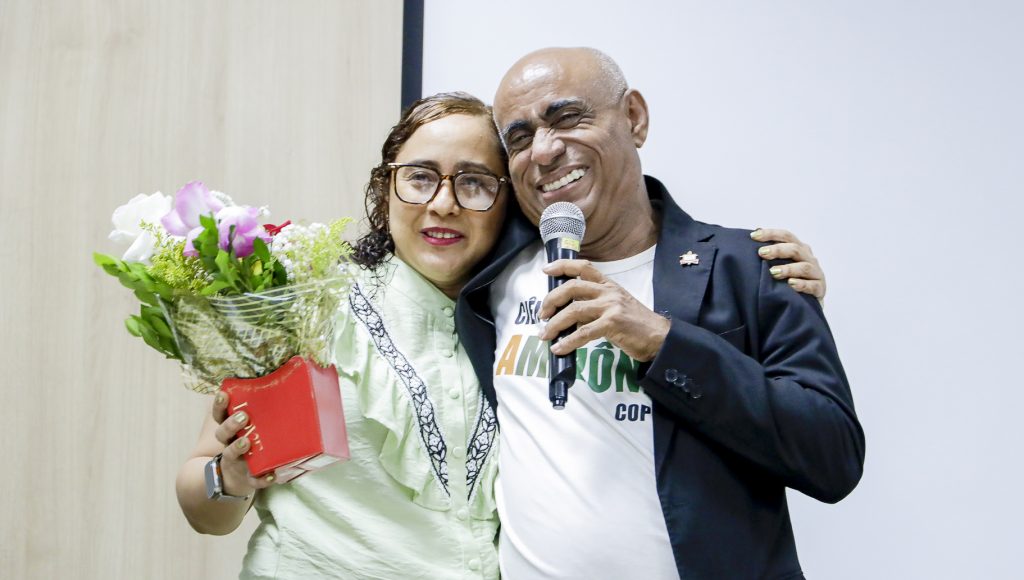 Fotografia da servidora da UFPA Valda Andrade ao lado do reitor da UFPA, Gilmar Pereira da Silva. O reitor fala ao microfone e a servidora segura um vaso com flores.
