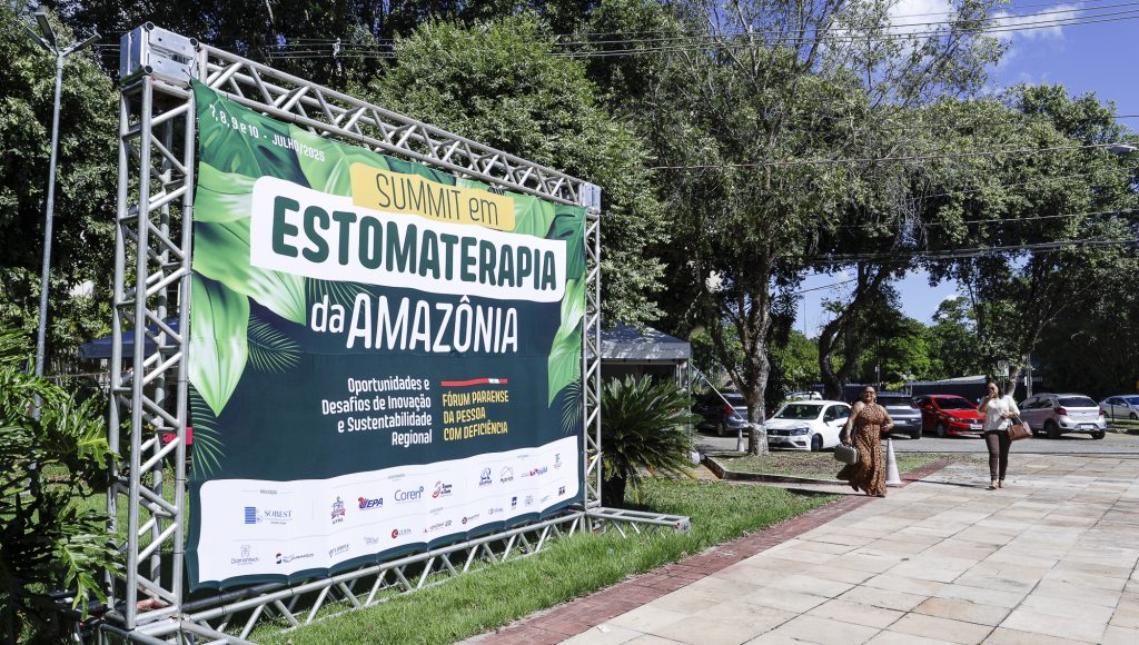 Fotografia registra um grande barnner disposto em armações de ferro. O banner apresenta o seguinte texto: Summit em Estomaterapia da Amazônia - OPORTUNIDADES E DESAFIOS DE INOCAÇÃO E SUSTENTABILIDADE REGIONAL - Fórum Paraense da Pessoa com Deficiência.