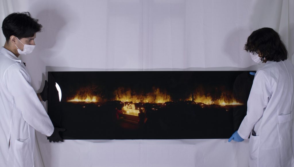 Fotografia registra duas pessoas segurando um quadro retangular próximos a uma parede branca. O quadro, de moldura preta, retrata uma paisagem escura com destaque para pontos de fogo.
