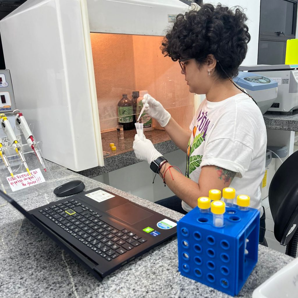Fotografia registra Viviane Firmino em ação dentro de um laboratório. Ela usa luvas plásticas, manuseia tubos de ensaio e dosadores. A frente da pesquisadora há um notebook.