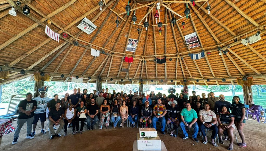 Fotografia posada de um grupo de pessoas. Eles estão em uma estrutura que lembram uma maloca, com chão de terra batida. No teto, é possível identificar cartazes e maracás pendurados no teto de palha.