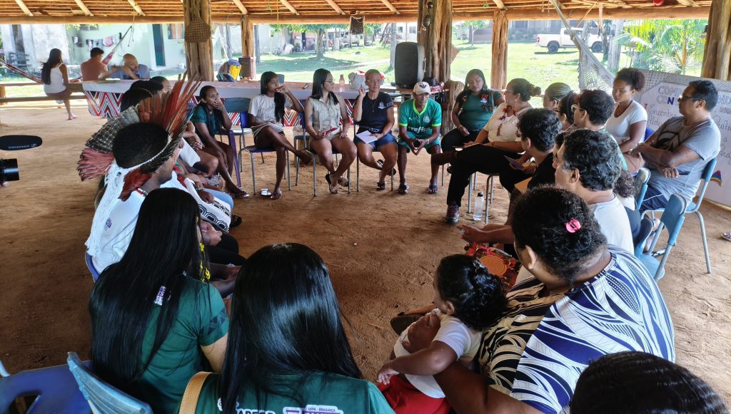 Fotografia registra uma roda de conversa em um território indígena. Um grupo de pessoas está sentado em cadeiras disposta em um círculo, há adultos e crianças.