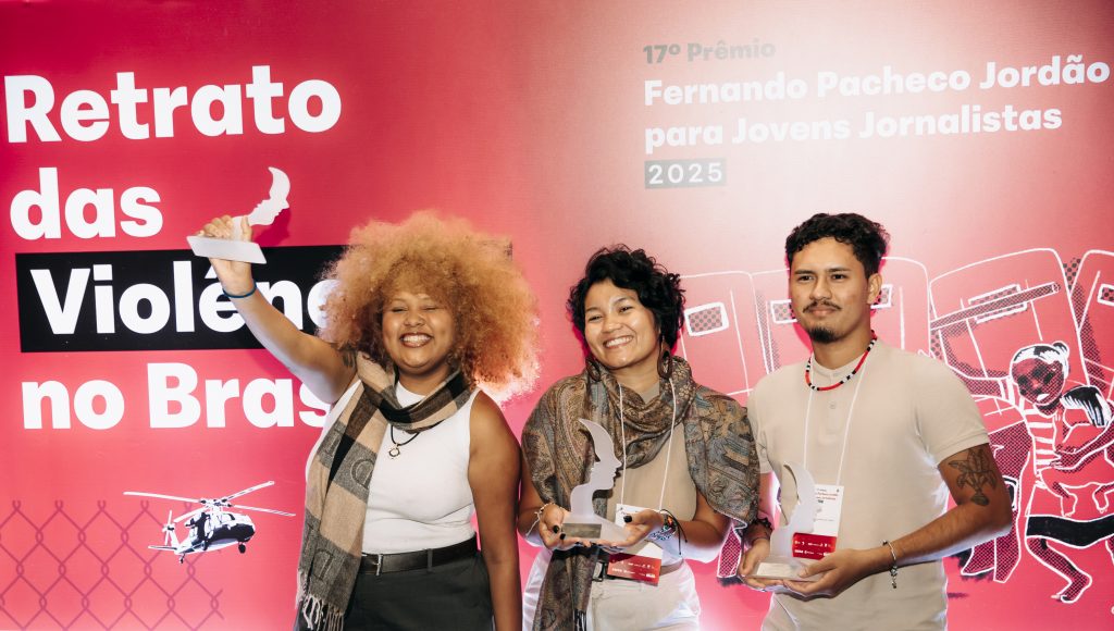 Fotografia registra três pessoas a frente de um painel com o seguinte texto: RETRATOS DAS VIOLÊNCIAS NO BRASIL. Entre as pessoas, duas mulheres e um homem. Cada um segura um troféu.