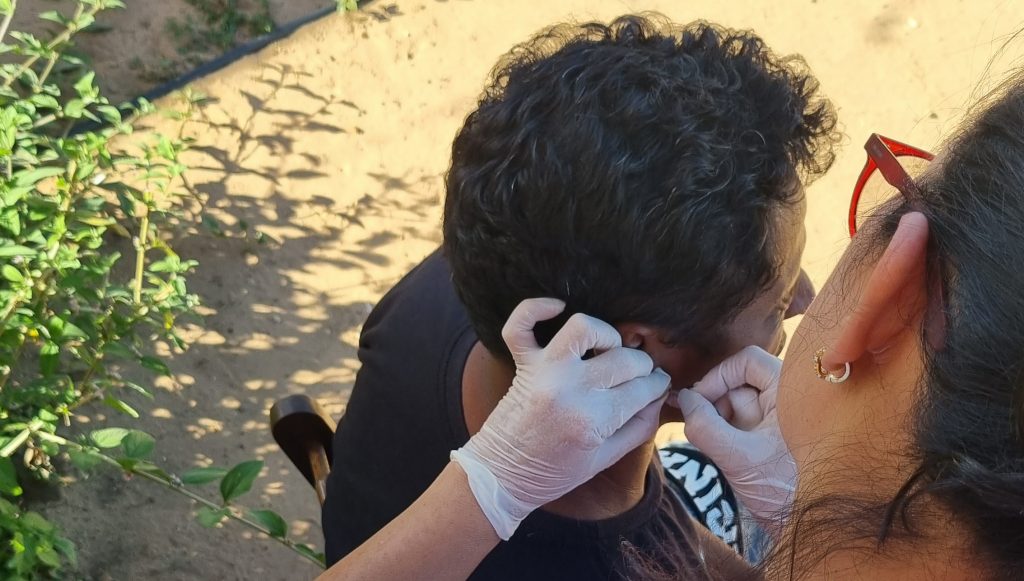 Fotografia registra duas pessoas. Uma esta sentada e é registrada de perfil, outra está de costa, podemos ver suas mãos com luvas decaratáveis manuseando algum aparelho na orelha da pessoa sentada.