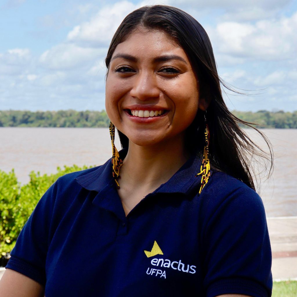 Fotografia posada da estudante Evelyn Mesquita. Ela tem cabelos pretos, lisos e longos. Ela usa blusa azul com logo da Enactus e brincos amarelos.