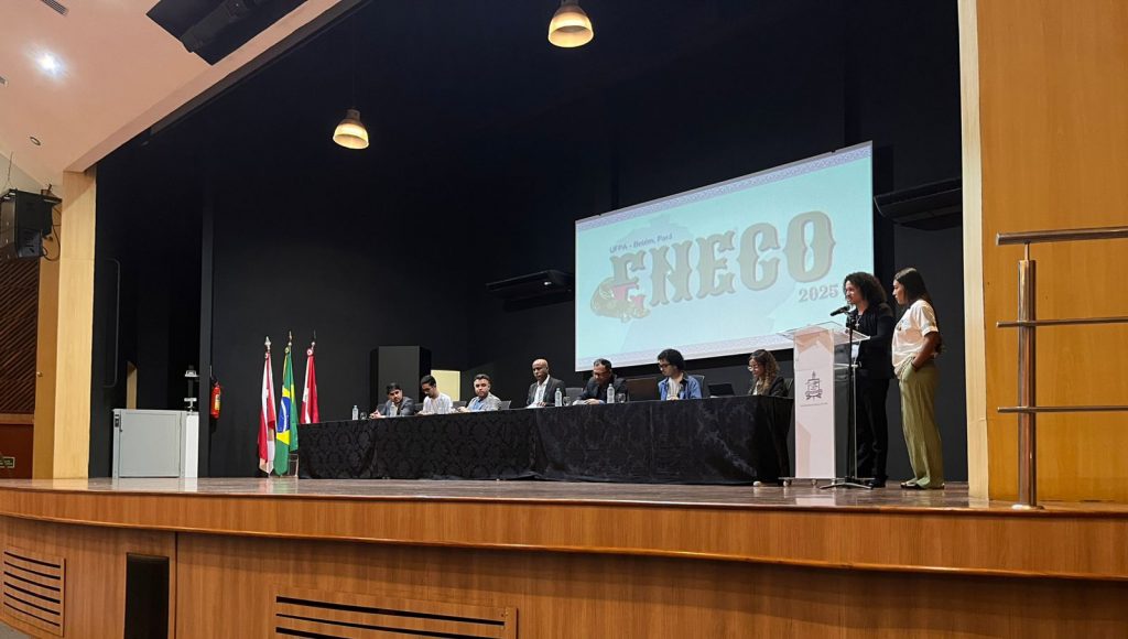 Fotografia registra o palco de uma evento. Há uma mesa com várias pessoas sentadas, a frente de um telão com a palavra "ENECO 2025".