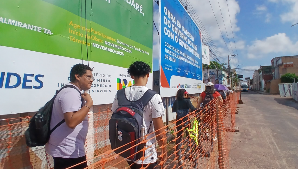 Fotografia registra um grupo de pessoas caminhando por uma área na lateral de uma pista. A área está cercada por material plástico laranja. É possível identificar grandes sinalizações de obras.