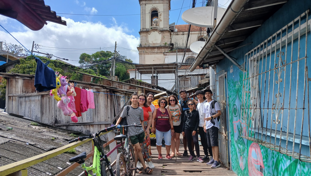 Fotografia posada de um grupo de pessoas, eles estão um lugar cercado por estrutaras (ponte e casa) de madeira. Ao fundo, é possível identificar parte de uma construção típica de igrejas.