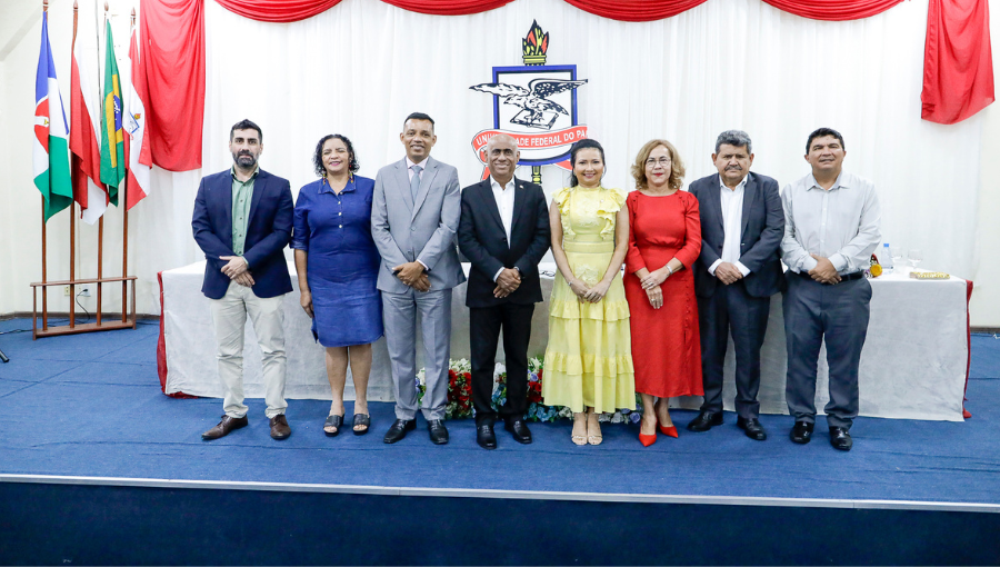 Fotografia posada das autoridades que compuseram a mesa da cerimônia da posse. As autoridades, homens e mulheres, estão de pé à frente de uma mesa retangular. À esquerda há um mastro com três bandeiras e, por detrás do grupo, vê-se o brasão da UFPA.