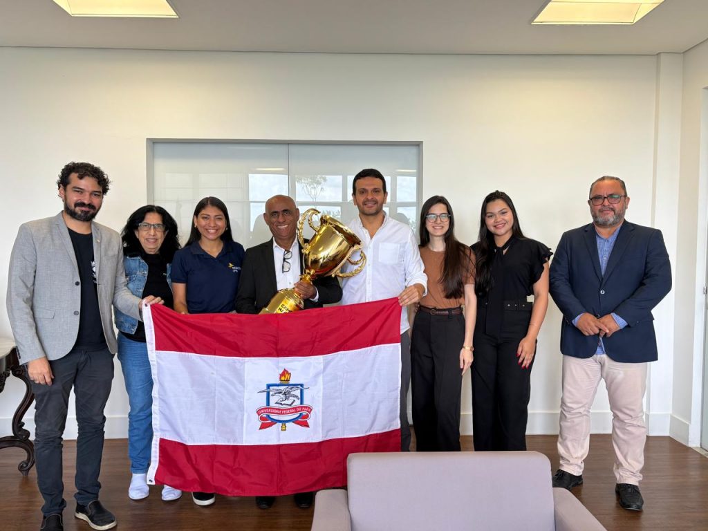 Fotografia mostra grupo de pessoas à frente de uma parede branca. O reitor da UFPA, homem negro, segura um troféu dourado. Os demais sustentam a bandeira da UFPA.
