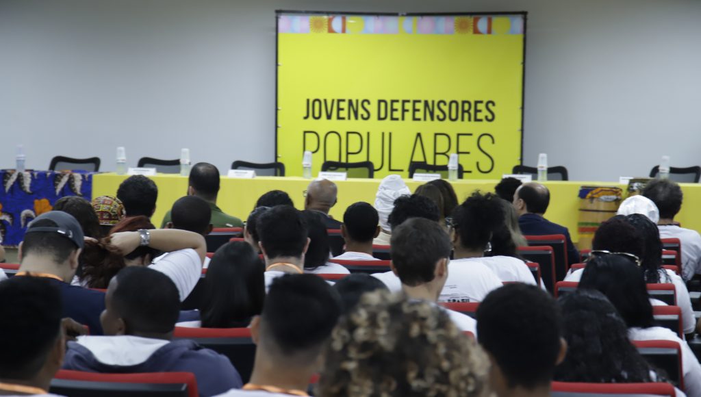 Fotografia registra a plateio de um auditório de costa. Ao fundo, é possível identificar um telão amarelo que destaca a mensagem: Projeto Jovens Defensores Populares.