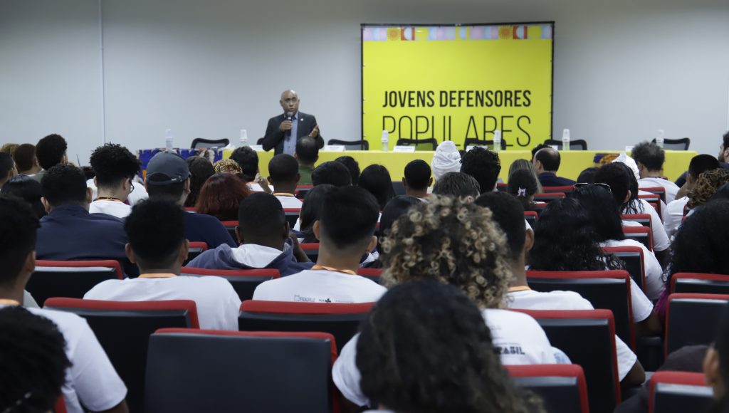 Fotografia registra a plateio de um auditório de costa. Ao fundo, é possível identificar um telão amarelo que destaca a mensagem: Projeto Jovens Defensores Populares. A frente do painel, o reitor da UFPA, Gilmar Pereira da Silva, fala ao microfone.