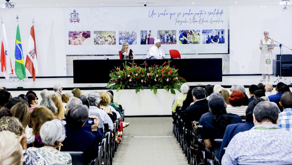 Fotografia de parte de um auditório. Ao fundo, em uma mesa, o reitor da UFPA. Na lateral, em um púlpito, a professora Terezinha fala ao microfone.