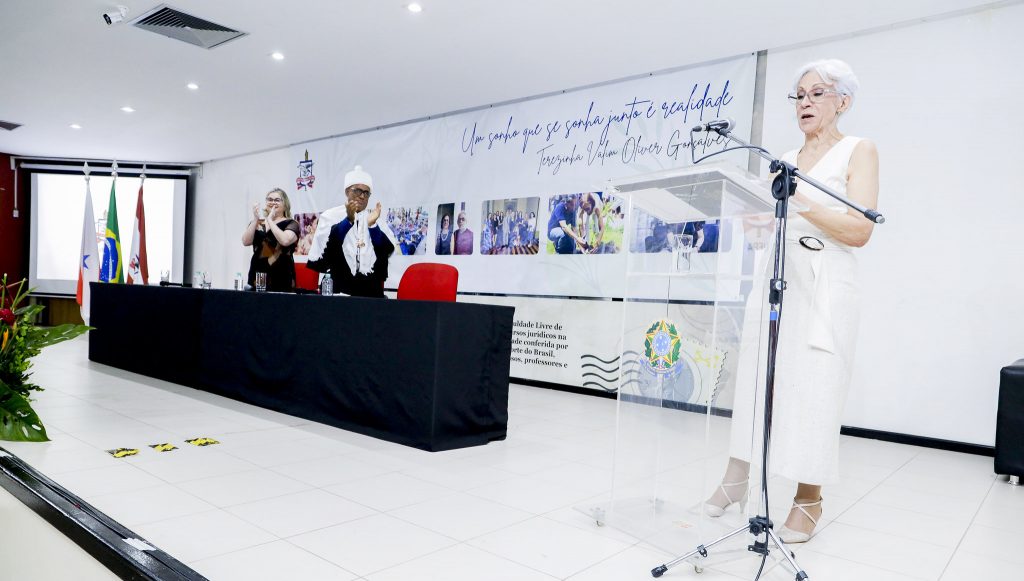Fotografia registra o momento que a professora Terezinha Valim fala ao microfone, em um púlpito, e é aplaudida pelo reitor da UFPA, Gilmar Pereira da Silva, e pela professora  Franci Sarmanho.