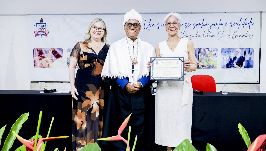 Fotografia posada da professora Terezinha Valim Oliver Gonçalves, com o reitor da UFPA, Gilmar Pereira da Silva, e a professora Franci Sarmanho. Terezinha segura uma placa.