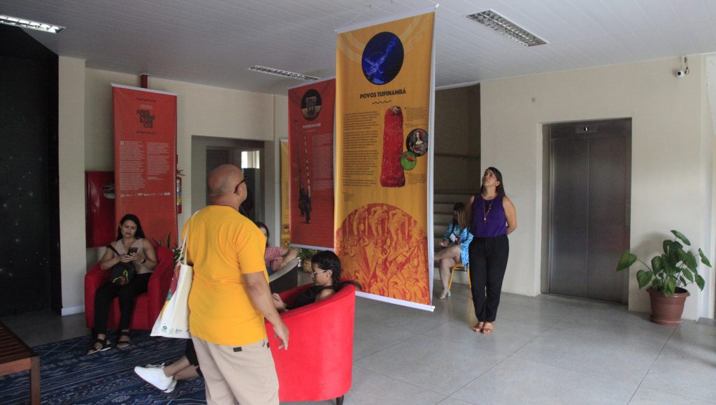 Fotografia de uma espaço fechado com vários banners pendurados do teto ao chão. Algumas pessoas observam os banners.