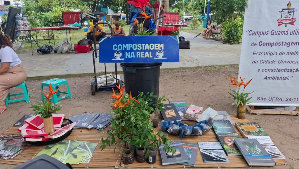 Fotografia de um espaço aberto. No chão, várias publicações e mudas de plantas estão dispostos sobre esteiras. Ao fundo, a placa "COMPOSTAGEM NA REAL".