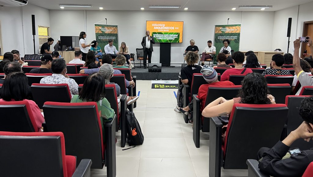 Fotografia de um auditório, com destaque para um grupo de pessoas no palco ao fundo. Em pé, no palco, o reitor da UFPA, Gilmar Pereira da Silva, fala ao microfone.