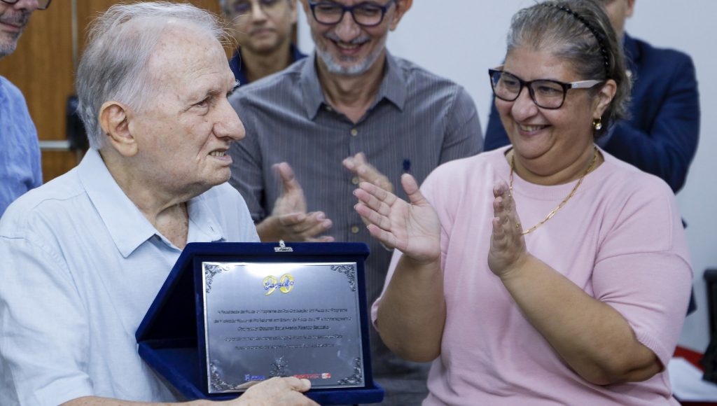 Fotografia registra o professor Bassalo, segurando uma placa comemorativa, sendo aplaudido por outras pessoas que estão no segundo plano da imagem.