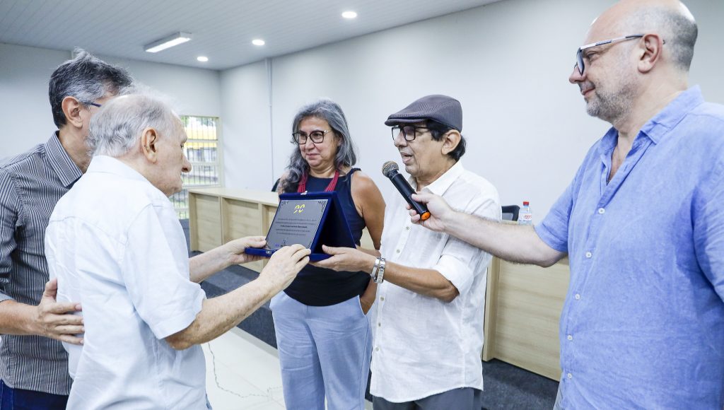 Fotografia registra o professor Bassalo recebendo um placa comemorativa das mãos de um grupo de docentes.