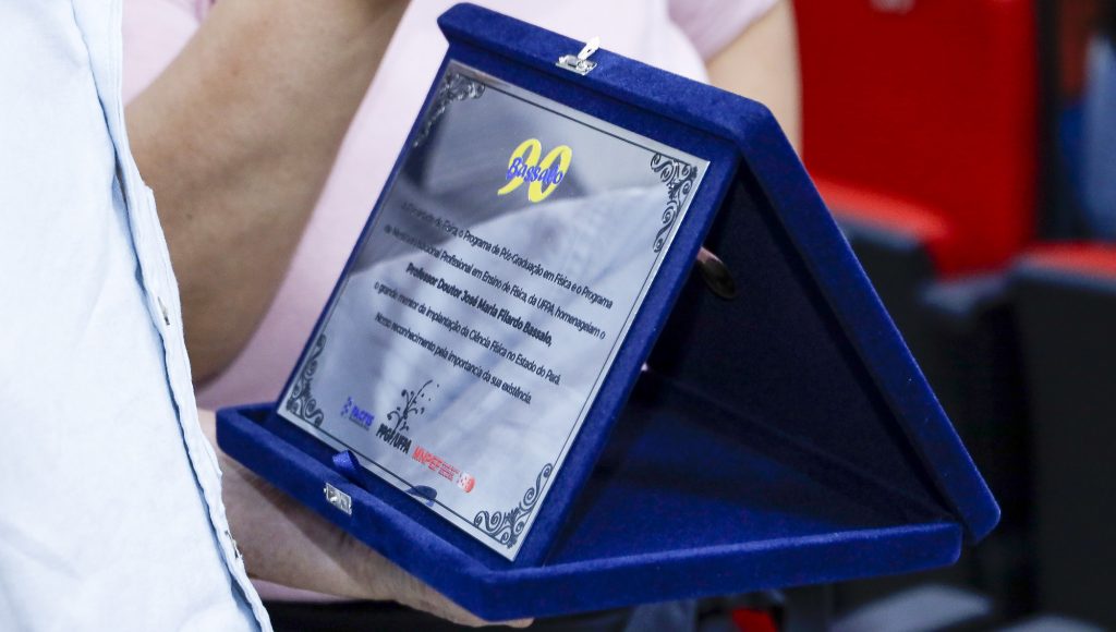 Fotografia registra uma placa de metal, dentro de um estojo azul, com a inscrição "Bassalo 90", seguida por um grande texto.