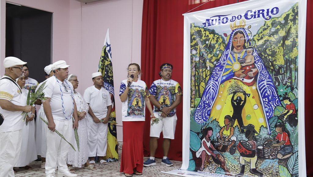 Fotografia registra a professora Inês Ribeiro, falando ao microfone, enquanto aponta para o cartaz do Auto do Círio 2025.