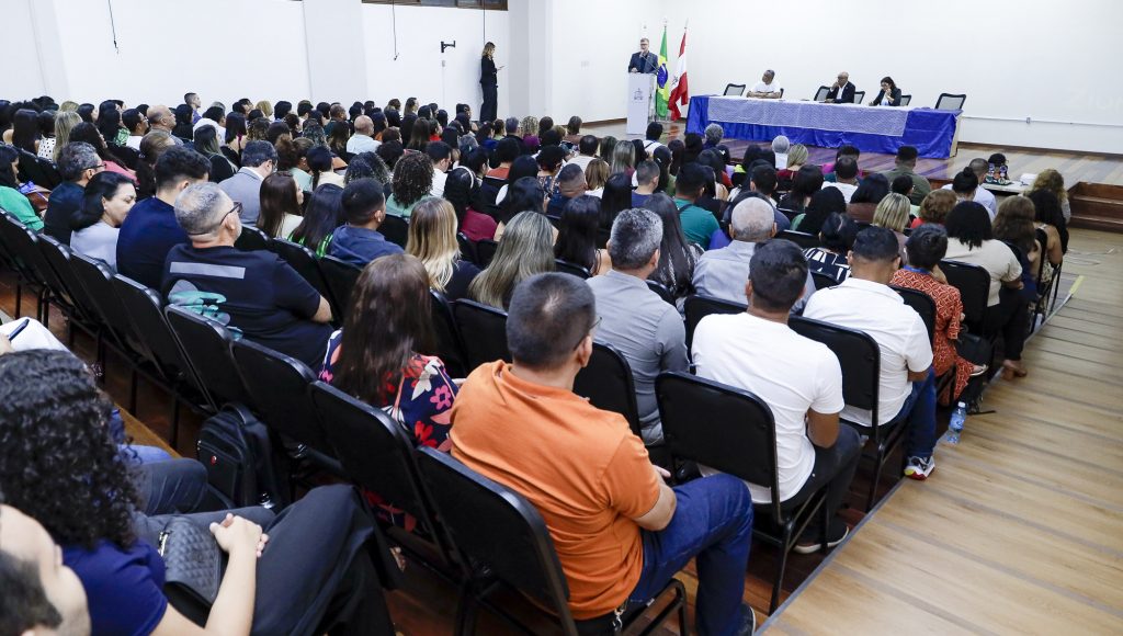 Fotografia registra a plateia de um evento. O auditório está lotado e, ao fundo, é possível ver a mesa do evento com três pessoas sentadas e uma quarta pessoa falando ao microfone.