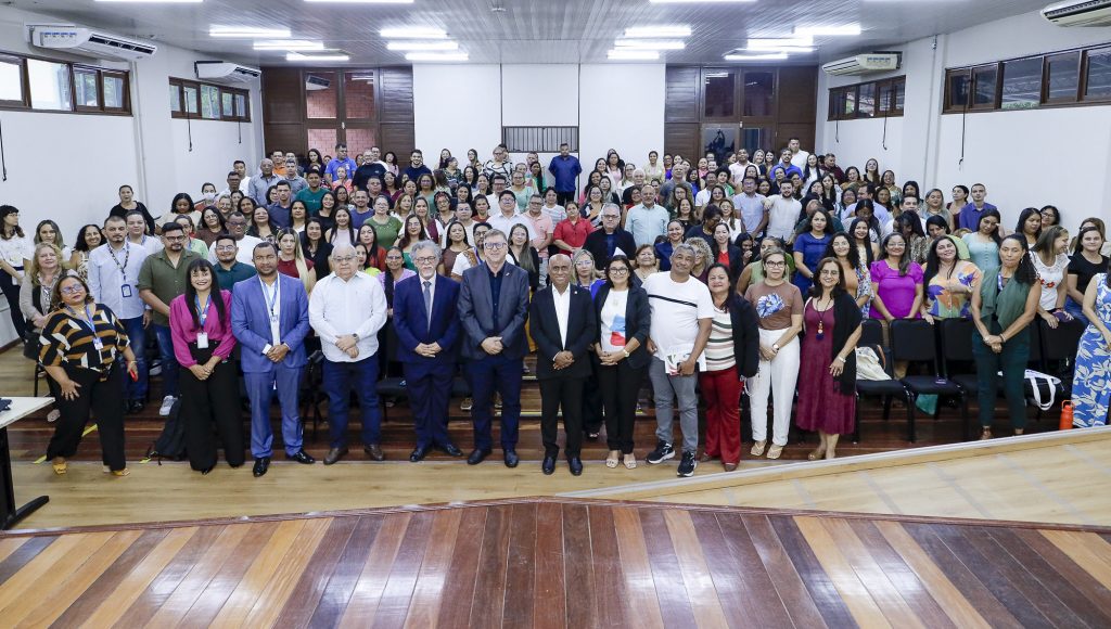 Fotografia posada de um grupo de um grande grupo de pessoas. Todos estão dentro de um auditório, em pé, olhando para a câmera. à frente do grupo estão o reitor da UFPA, Gilmar Pereira da Silva, e o secretário estadual de Assistência Social, Sistema Único de Assistência Social, Inocêncio Gasparim.