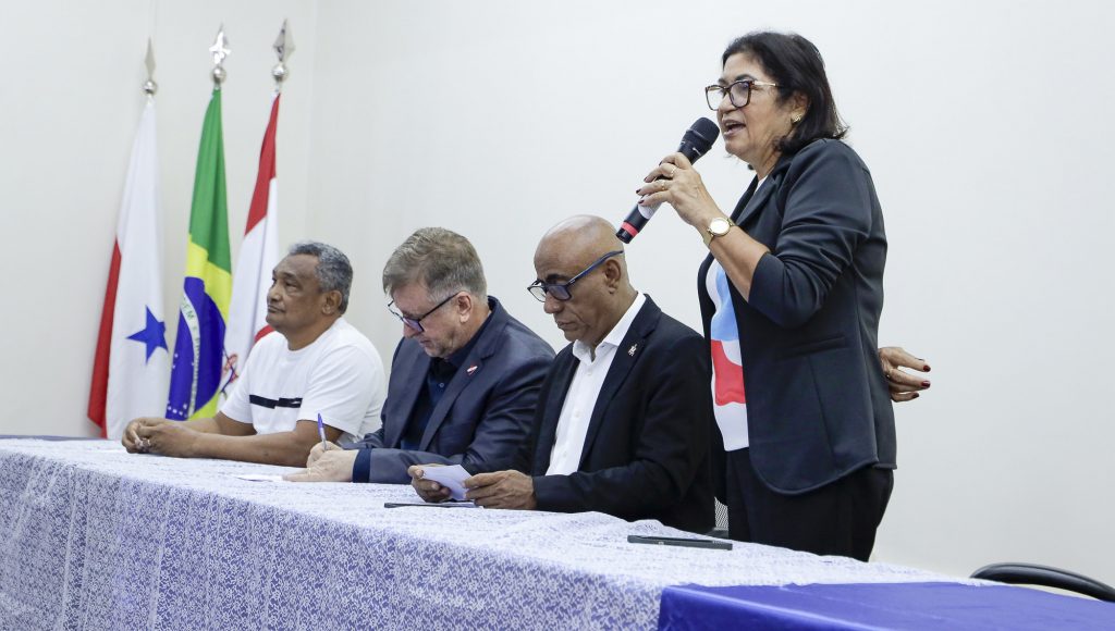 Fotografia registra a mesa do evento formada por quatro pessoas, sendo três homens e uma mulher. A mulher está em pé, falando ao microfone.