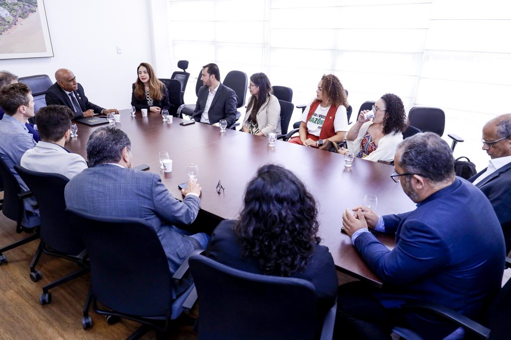 Fotografia de grupo de pessoas. Todos estão sentados ao redor de uma mesa de madeira.
