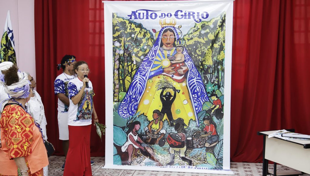 Fotografia registra a professora Inês Ribeiro, falando ao microfone, enquanto aponta para o cartaz do Auto do Círio 2025.