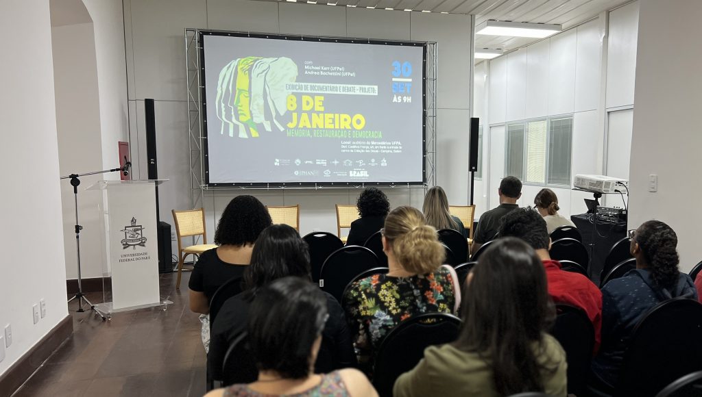 Fotografia registra uma plateia de costa olhando para uma tela onde está projetada a capa de um filme, com o seguinte texto: EXIBIÇÃO DE DOCUMENTÁRIO E DEBATE - 8 DE JANEIRO, MEMÓRIA, RESTAURAÇÃO E DEMOCRACIA - 30 DE SETEMBRO, ÀS 9H.