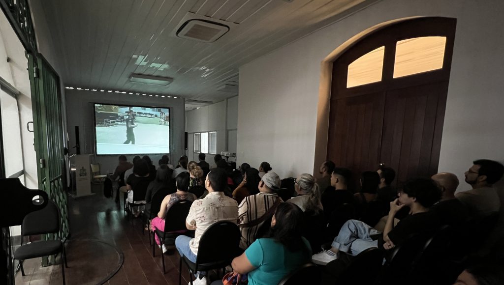 fotografia REGISTRA UMA GRANDE PLATEIA, DE COSTA, ATENTA A UM TELÃO QUE REPRODUZ IMAGENS DE UMA FILME.