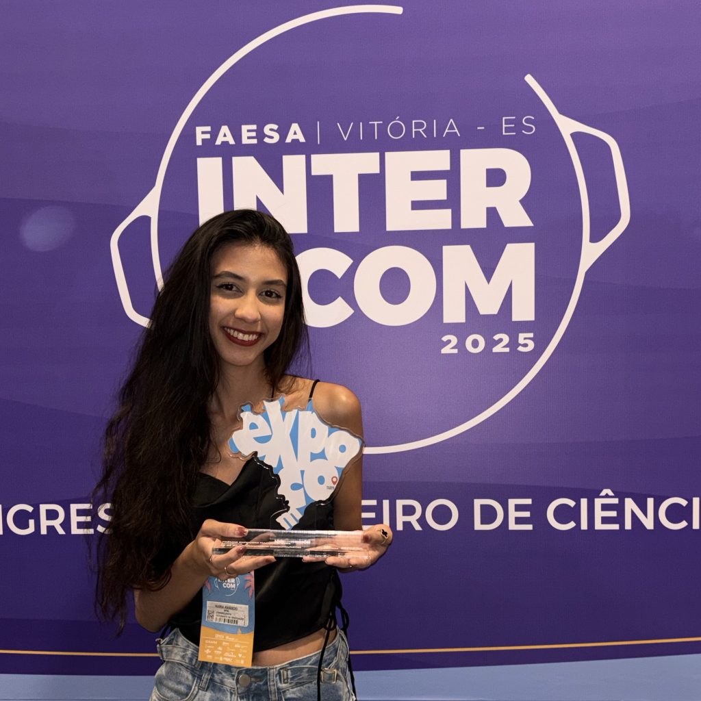 Fotografia posada da estudante Luiza Amâncio. Ela está a frente de um banner escrito "intercom 2025" e segura um troféu no formato do mapa do Brasil.